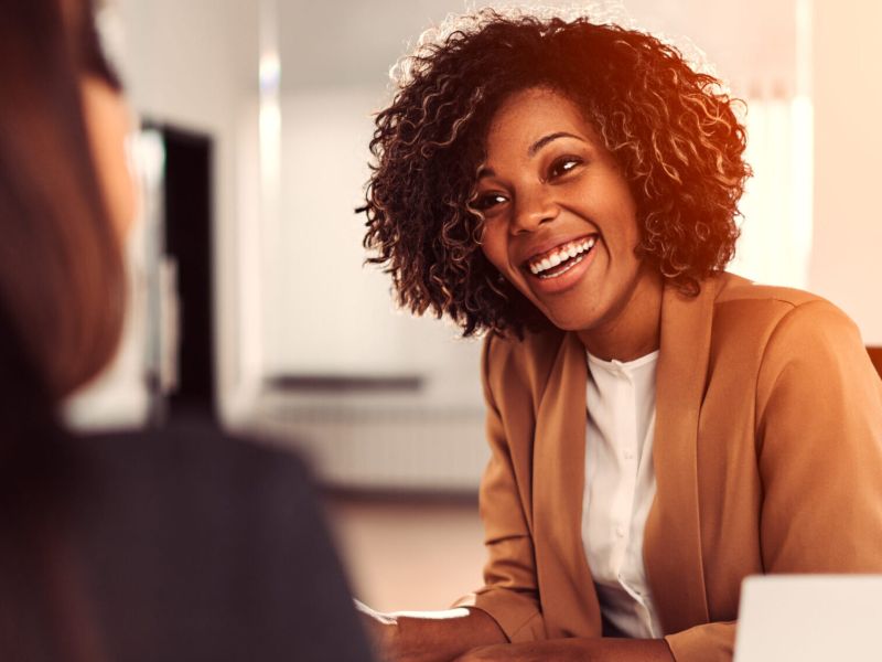 Business woman in a tan jacket is smiling and engaging with a potential client.