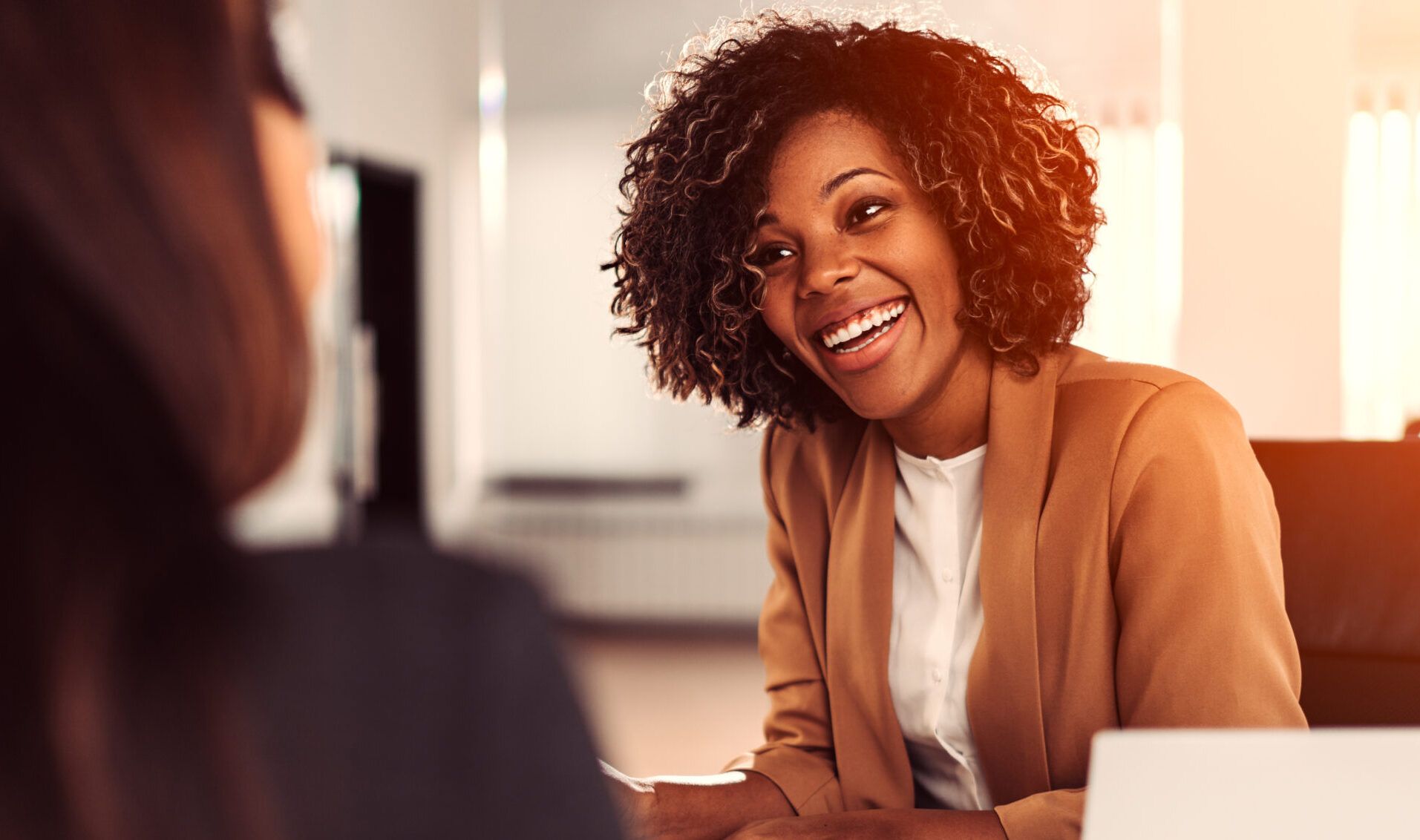 Business woman in a tan jacket is smiling and engaging with a potential client.